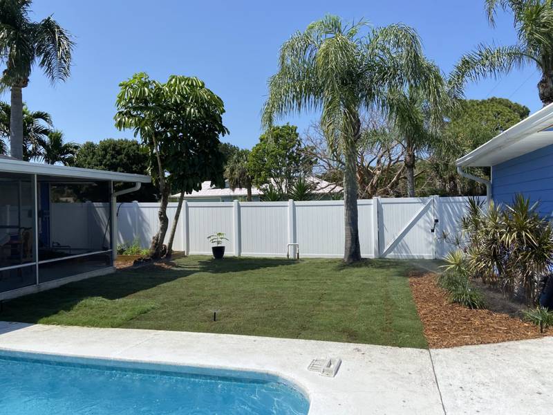 New sod between a pool deck and a white vinyl fence, with palms beyond