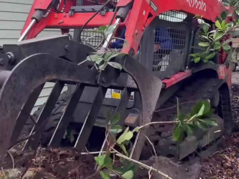 Compact track loader with a grapple lifting cut branches beside a house
