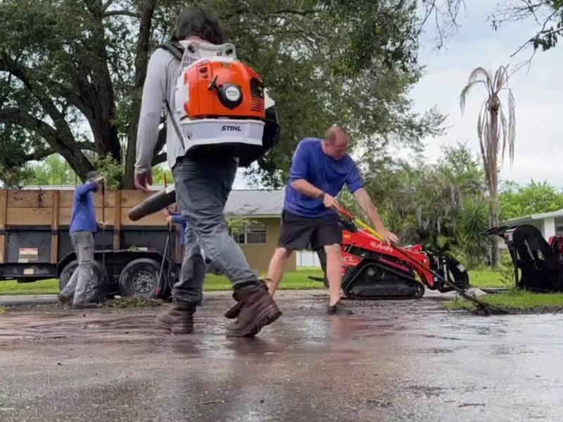 Crew blowing storm debris off a wet street next to the trailer and track loader